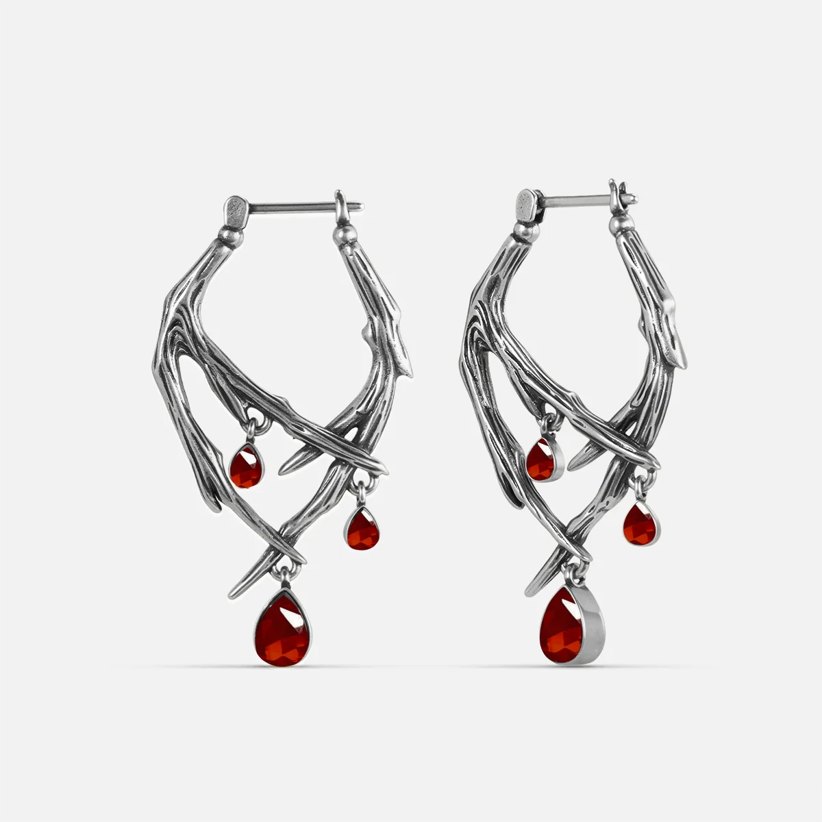 Silver-tone thorn-shaped hoop earrings pair with dangling red teardrop crystals, front view on white background